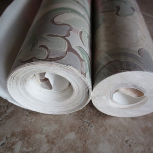 May include: Two rolls of wallpaper, one partially unrolled, showing a repeating pattern of green, brown, and grey floral designs. The rolls are tightly wound, revealing the edges of the paper.