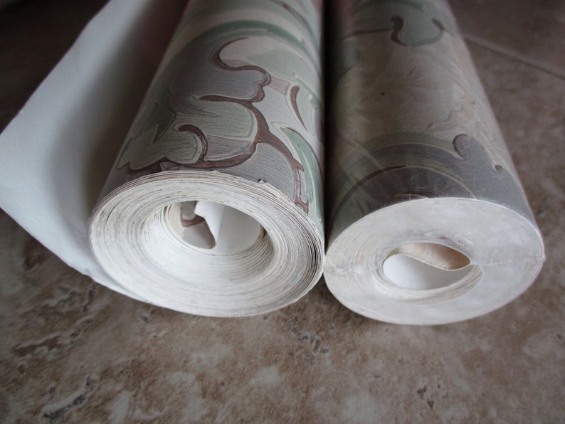 May include: Two rolls of wallpaper, one partially unrolled, showing a repeating pattern of green, brown, and grey floral designs. The rolls are tightly wound, revealing the edges of the paper.