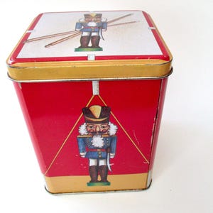 May include: A red metal tin with a gold rim and a white top. The tin features illustrations of a nutcracker soldier. The nutcracker is wearing a blue jacket, white collar, and a black hat. The tin is a trapezoid shape.