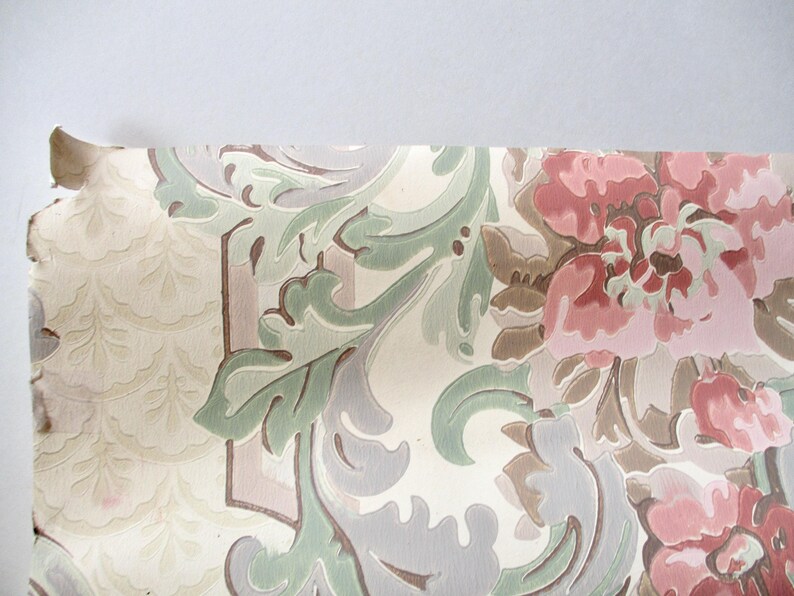 May include: A fragment of vintage wallpaper with a floral and scroll design. The pattern features large pink and brown flowers, green leaves, and grey scrollwork on a cream background. The edge of the paper is torn.