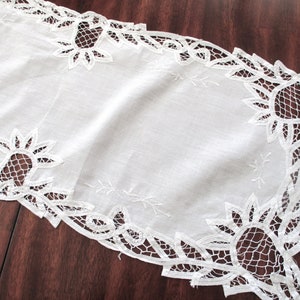 May include: White linen table runner with intricate lace detailing. The runner features a delicate floral and geometric lace design along the edges, adding a touch of elegance. Perfect for adding a vintage touch to any dining table.