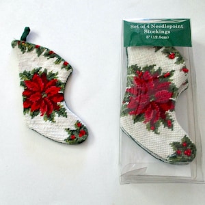May include: Needlepoint Christmas stocking with a poinsettia design. The stocking is white with a red poinsettia and green leaves. The package reads "Set of 4 Needlepoint Stockings 5" (12.5cm)".