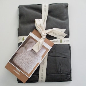 May include: A folded, dark gray bedding set tied with a cream ribbon printed with "west elm". A product tag shows a close-up of a white patterned pillowcase. The tag reads "SHAM - STANDARD".