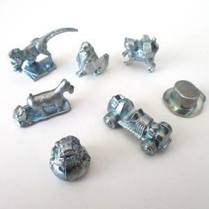 May include: A collection of silver-colored game pieces, including a dinosaur, dog, cat, goat, race car, top hat, and a small, round object. The pieces are arranged on a white surface.