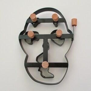 May include: A black metal cookie cutter in the shape of a face, with wooden pegs. The cutter has a handle and is designed to cut out multiple shapes at once. The cutter is approximately 7 inches tall and 6 inches wide.