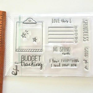 May include: A clear stamp set for budget planning, featuring phrases and images. Includes stamps with text such as "Budget Tracking," "Love this!," and "No Spend Month." Also features star and heart designs.