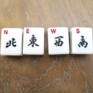 May include: Four white Mahjong tiles spelling "NEWS". Each tile has a letter in red and a black Chinese character. The tiles are arranged on a wooden surface.
