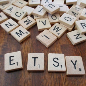 May include: Wooden Scrabble tiles spelling out the word "ETSY" on a wooden surface, with additional tiles scattered around. The letters are black, and the tiles are beige.