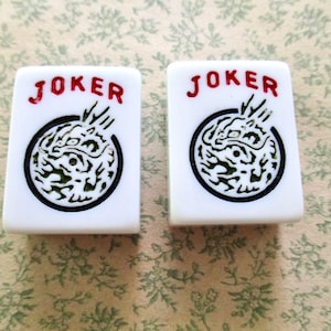 May include: Two white Mahjong tiles with the word "JOKER" in red at the top. Each tile features a black circle with a green and white dragon design. The tiles are rectangular with rounded corners.