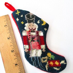 May include: Needlepoint Christmas stocking featuring a nutcracker soldier in red and white uniform against a navy blue background. The stocking is approximately 4 inches long, with a red loop for hanging.
