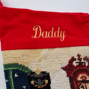 May include: A Christmas stocking with a red velvet top embroidered with the word "Daddy" in gold script. The lower portion features a needlepoint design with houses in shades of green, black, and red.