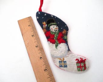 May include: A miniature Christmas stocking ornament, featuring a snowman in a red coat and green scarf against a dark blue starry background. The stocking is approximately 4 inches long and has a red hanging loop.