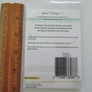 May include: A packet of Pinkfresh Studio clear stamps, with instructions and a barcode. The packaging includes text that reads "Good job" and "Made in the USA". A wooden ruler is placed next to the packet.