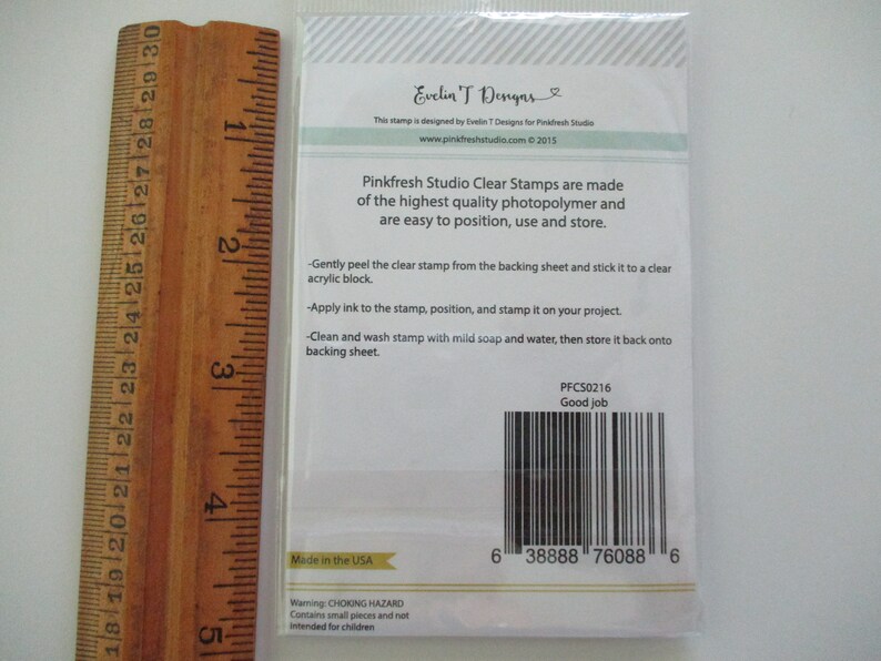 May include: A packet of Pinkfresh Studio clear stamps, with instructions and a barcode. The packaging includes text that reads "Good job" and "Made in the USA". A wooden ruler is placed next to the packet.
