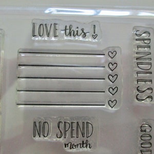 May include: Clear acrylic stamps with black text. The stamps include phrases like "LOVE this!", "SPENDLESS", and "NO SPEND month". Other stamps feature a checklist design with hearts.