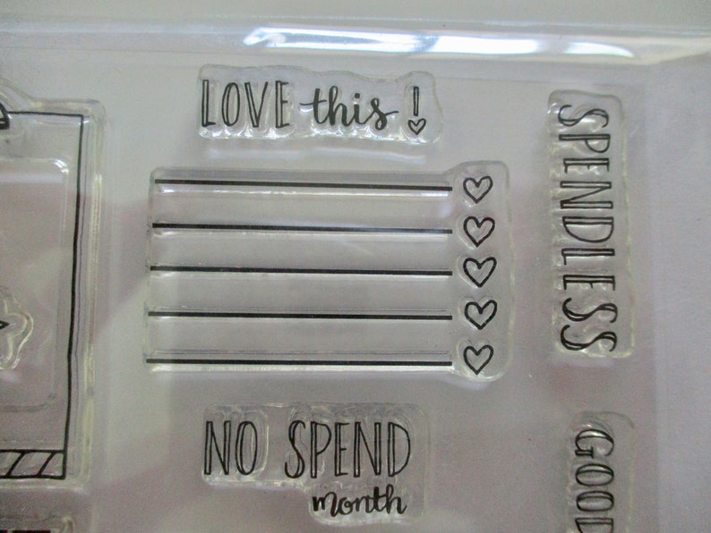 May include: Clear acrylic stamps with black text. The stamps include phrases like "LOVE this!", "SPENDLESS", and "NO SPEND month". Other stamps feature a checklist design with hearts.