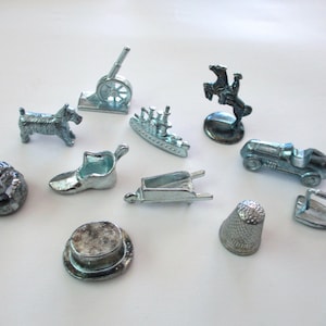 May include: A collection of silver-colored Monopoly game tokens, including a dog, cannon, ship, race car, horse and rider, shoe, wheelbarrow, hat, thimble, and iron. These classic game pieces are displayed on a white surface.
