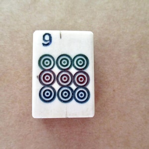 May include: A rectangular off-white mahjong tile featuring the number 9 in blue at the top. Below the number, there is a grid of nine circular symbols arranged in three rows, with the top row in green, the middle row in red, and the bottom row in blue.