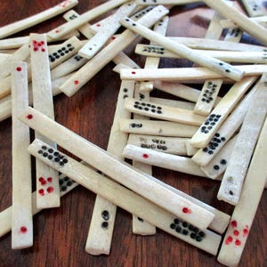 May include: A collection of ivory-colored dominoes with black and red dot markings. The dominoes are scattered on a dark wooden surface, showcasing the game pieces' varied dot patterns and shapes.