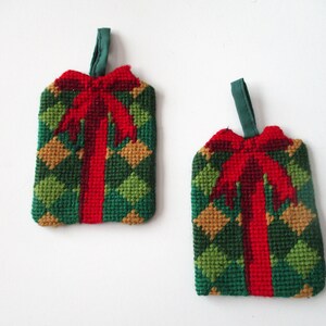 May include: Two handmade Christmas ornaments shaped like gift boxes. The ornaments feature a green and gold diamond pattern with a red bow and ribbon. Each ornament has a green loop for hanging.