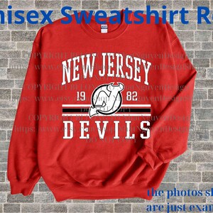 May include: A red unisex sweatshirt with the text "NEW JERSEY DEVILS" in white. The design includes a logo with a devil and the numbers "19" and "82". The sweatshirt is a crew neck style.