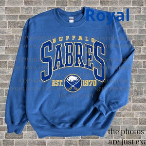 May include: Royal blue sweatshirt with a gold and blue Buffalo Sabres logo. The logo includes the team name, "Buffalo Sabres", and the text "EST. 1970".