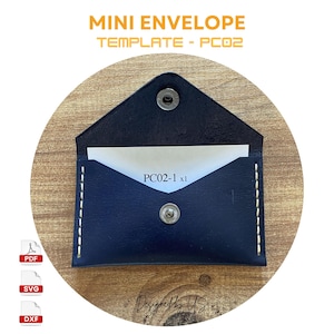 Puede incluir: Un mini sobre de cuero azul con detalles de costura blanca y dos broches de presión plateados. El sobre está sobre un fondo de madera. El texto "PC02-1 x1" está impreso en el interior del sobre.