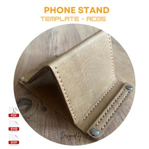 Puede incluir: Un soporte para teléfono de cuero marrón claro con costuras y remaches plateados. El soporte está plegado y colocado sobre una superficie de madera. El texto "PHONE STAND TEMPLATE - AC05" está en la parte superior de la imagen. El texto "PDF, SVG, DXF" está en la esquina inferior izquierda de la imagen. El texto "Designed by U.S." está en la esquina inferior derecha de la imagen.
