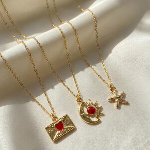 Puede incluir: Collares dorados con colgantes únicos. Uno presenta un sobre con un corazón rojo, otro un sol y una luna creciente con un centro rojo, y el último una estrella de mar. Las cadenas son delicadas y los colgantes están detallados.