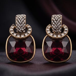 May include: A pair of drop earrings featuring large, square-cut deep red crystals set in antique gold-toned metal. Each earring has a circular accent and a rectangular top piece, both encrusted with small, clear white rhinestones.