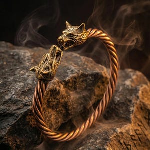 Può includere: Un braccialetto color bronzo con un design a corda intrecciata. Il braccialetto presenta due elementi decorativi a forma di testa di lupo rivolti l'uno verso l'altro. Il braccialetto è esposto su una superficie rocciosa scura e strutturata, con uno sfondo fumoso.