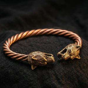 May include: A copper-colored bracelet with a twisted band and animal head terminals. The bracelet is made of metal and rests on a dark, textured surface. The animal heads are detailed with textured fur.