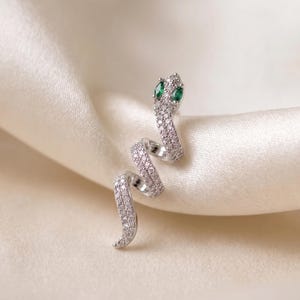 Könnte beinhalten: Ein silberfarbener Ohrring in Schlangenform mit einem gewundenen Körper, der mit zahlreichen kleinen, klaren Kristallen besetzt ist. Der Kopf der Schlange ist mit zwei marquise-geschliffenen grünen Steinen als Augen akzentuiert, was für ein detailliertes Aussehen sorgt.