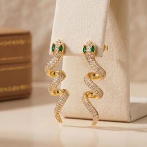 Puede incluir: Un par de pendientes dorados con forma de serpiente, con un cuerpo en espiral incrustado de pequeñas piedras transparentes y dos pequeñas piedras verdes que sirven de ojos. Los pendientes tienen cierre de presión y un acabado pulido.