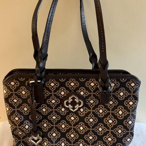 May include: A rectangular handbag with a geometric pattern in black, brown, and white. The bag has two braided brown handles and a silver-toned decorative clasp. A small charm hangs from the side. The bag is approximately 12 inches wide.