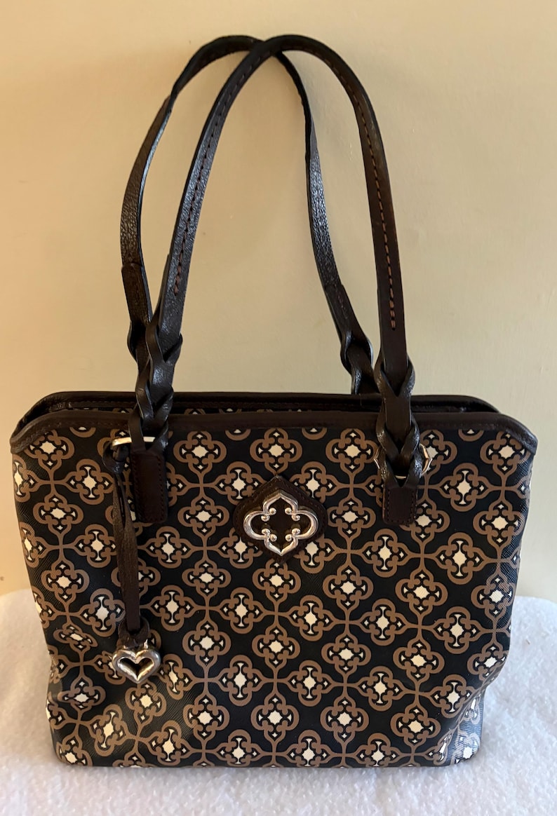 May include: A rectangular handbag with a geometric pattern in black, brown, and white. The bag has two braided brown handles and a silver-toned decorative clasp. A small charm hangs from the side. The bag is approximately 12 inches wide.