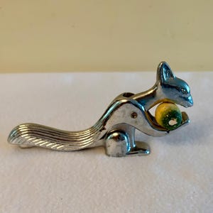 May include: A silver-toned metal nutcracker shaped like a squirrel with a long, textured tail. The squirrel figure is depicted holding a small, round, yellow and green nut between its front paws and its head, featuring a polished metallic finish.