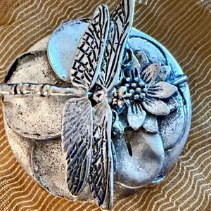 May include: A silver-toned metal dragonfly brooch. The brooch features a detailed dragonfly with textured wings and a flower. The dragonfly is positioned over a circular base with leaf and petal details. The brooch has an antique finish.