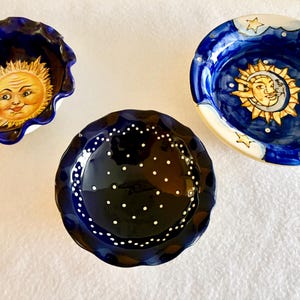 May include: Three ceramic bowls with scalloped edges. One bowl features a yellow sun face on a deep blue background. Another bowl displays a yellow sun and crescent moon face on a blue sky. The third bowl is dark blue with white dots.