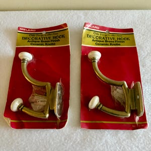 May include: Two decorative hooks from the Olde Forge Collection. Each hook has an antique brass finish and a white ceramic knob. The hooks are mounted on red cardboard packaging with the "MELARD" logo and product information.