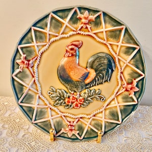 May include: Decorative ceramic plate featuring a rooster design. The plate has a cream-colored center with a detailed rooster illustration in shades of orange, blue, and green. The border is green with a star and rope design, and pink floral accents.