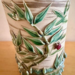 May include: A cylindrical ceramic vase with a cream-colored base featuring horizontal brown brushstrokes. The exterior is decorated with a raised, three-dimensional green bamboo stalk and leaf design, accented by a small, glossy red ladybug figurine.