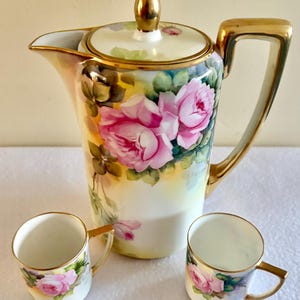 May include: A porcelain coffee pot with a lid and two matching cups, all featuring hand-painted pink roses, green leaves, and gold trim. The coffee pot has a gold handle, a gold spout, and a gold finial on the lid, while the cups have gold handles.