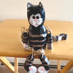 以下が含まれることがあります： 黒とグレーの縞模様の体、白いひげ、ピンクの鼻を持つ木製の猫の置物。 猫はぶら下がっている足と腕、縞模様の尾を持っています。 置物は木製のテーブルの上にあります。