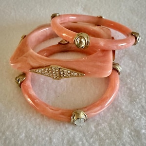 Puede incluir: Un conjunto de tres brazaletes de resina veteada en color melocotón. Uno de los brazaletes es cuadrado con un detalle central en forma de diamante de metal dorado incrustado con pedrería. Los otros dos brazaletes redondos tienen apliques de metal dorado.
