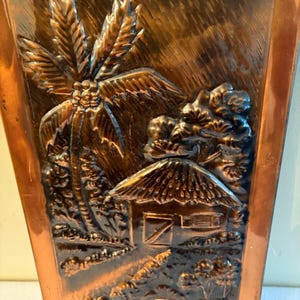 May include: A rectangular copper wall plaque featuring a relief design of a tropical scene. The embossed artwork depicts a palm tree, a small hut with a thatched roof, and a barrel in the foreground. The metal surface has a polished finish.