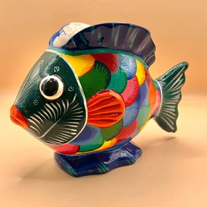 Pode incluir: Uma colorida figura de peixe em cerâmica com um design detalhado. O peixe apresenta uma cabeça verde escura, uma boca vermelha e um corpo adornado com tons vibrantes de azul, verde, amarelo e vermelho. O peixe tem uma cauda verde escura e está sobre uma base azul.