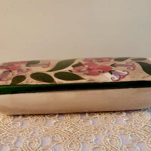 May include: Rectangular ceramic trinket box with a cream-colored base and a lid decorated with pink and purple floral designs and green leaves. The lid is outlined with a dark green border. The box is resting on a lace tablecloth.
