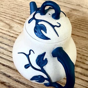 May include: A cream-colored ceramic teapot featuring a dark blue vine and leaf pattern. The teapot has a rounded body, a curved handle, a small spout, and a matching lid with a loop handle, all decorated with the same dark blue botanical motif.