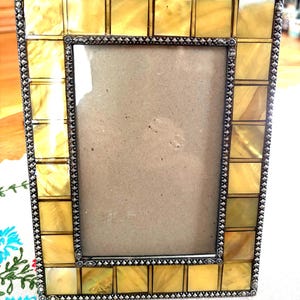May include: A rectangular picture frame featuring a border of square, yellow, iridescent tiles. The frame is edged with a dark, textured metal trim that has a repeating studded pattern along the outer and inner borders of the frame.